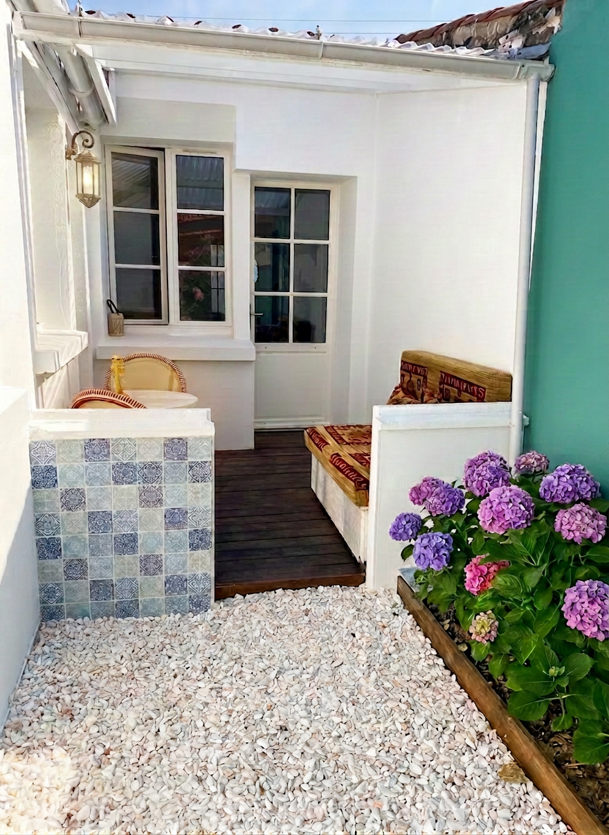 Remove the blue and white ceramic flower pot on the left side of the image, replacing it with the white gravel ground that surrounds it. Keep everything else exactly the same: the tiled wall, the wooden deck, the outdoor seating with cushions, the hydrangeas on the right, the windows, the lantern, and the turquoise wall.